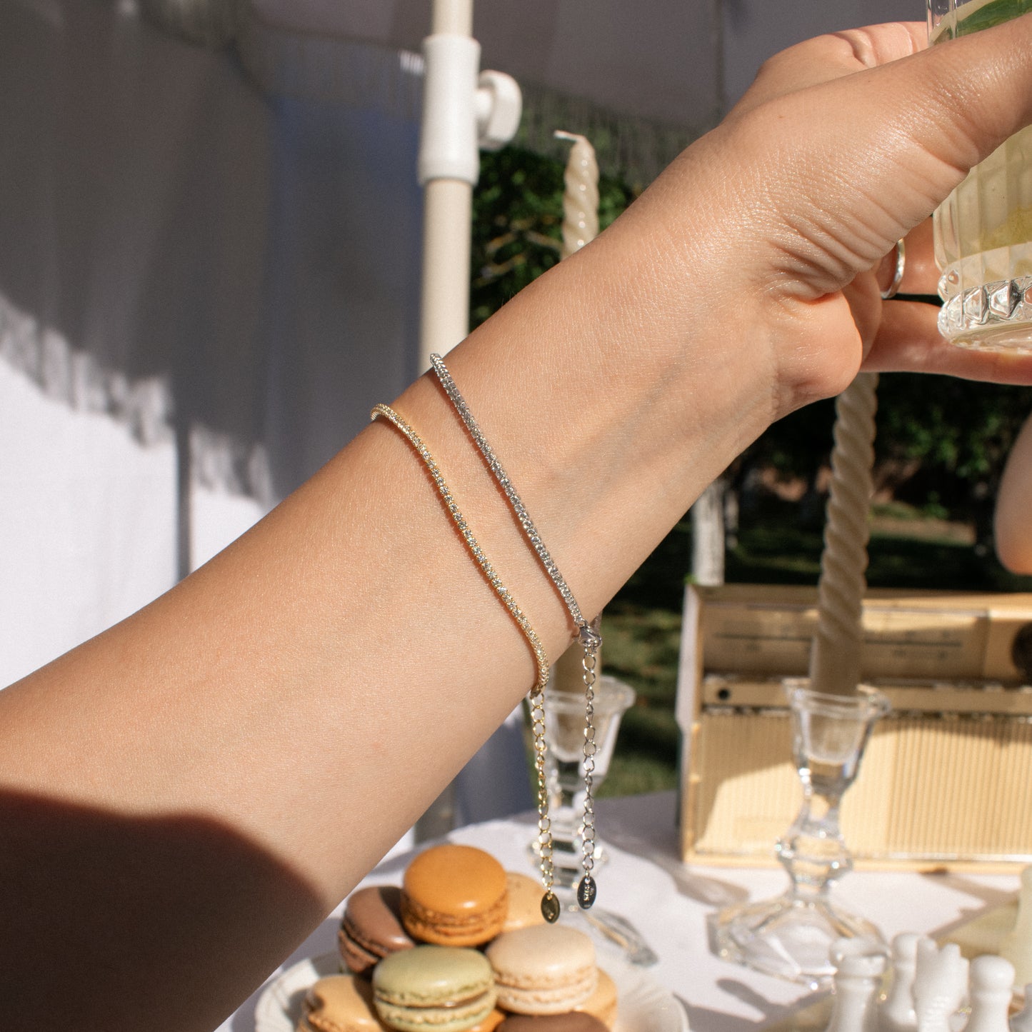 A close-up lifestyle shot of a person's arm wearing two sterling silver tennis bracelets, one gold plated and one silver, in an outdoor setting with macarons and glassware in the background.