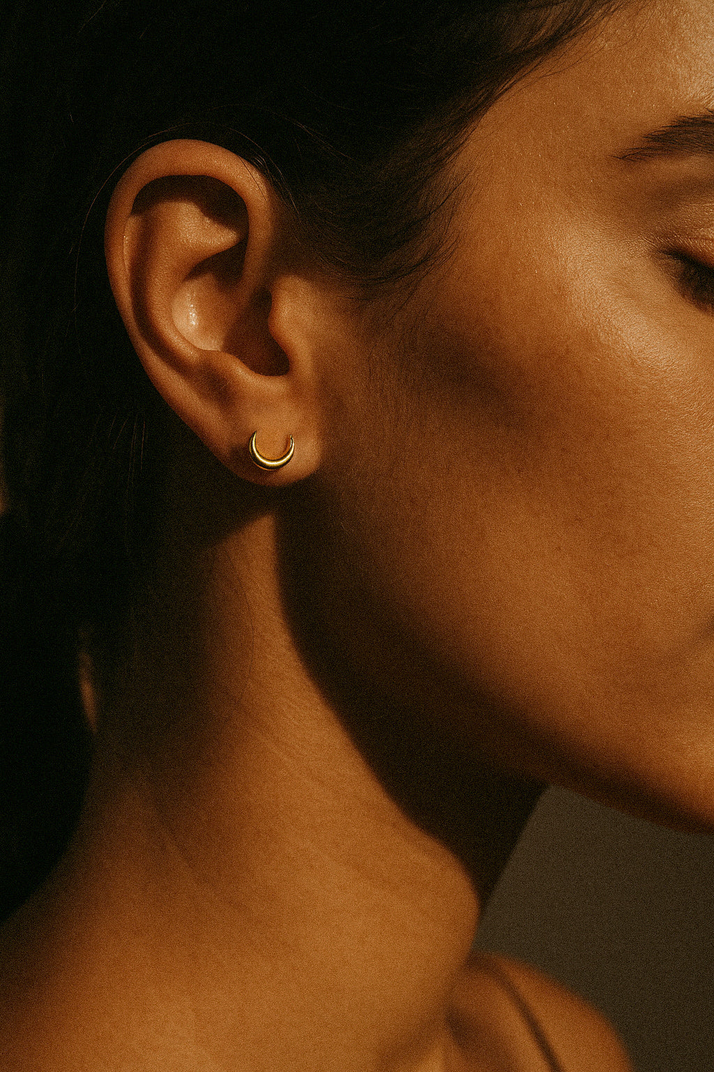 Prim-plan al urechii unei persoane cu design de semiluna, din aur solid de 14k. Close-up of a person's ear with a 14k solid gold earring with crescent moon design.