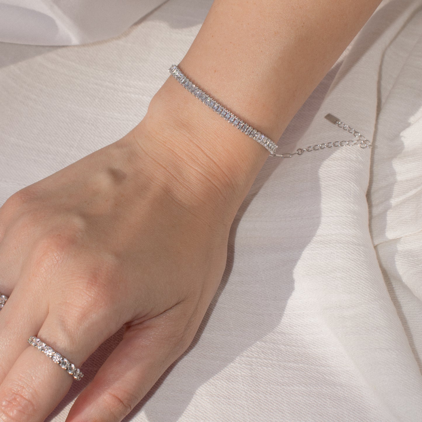 A close-up of a person's hand and wrist wearing a delicate silver-toned tennis bracelet with zirconia stones in emerald cut, paired with a matching stone band ring, resting on white fabric.