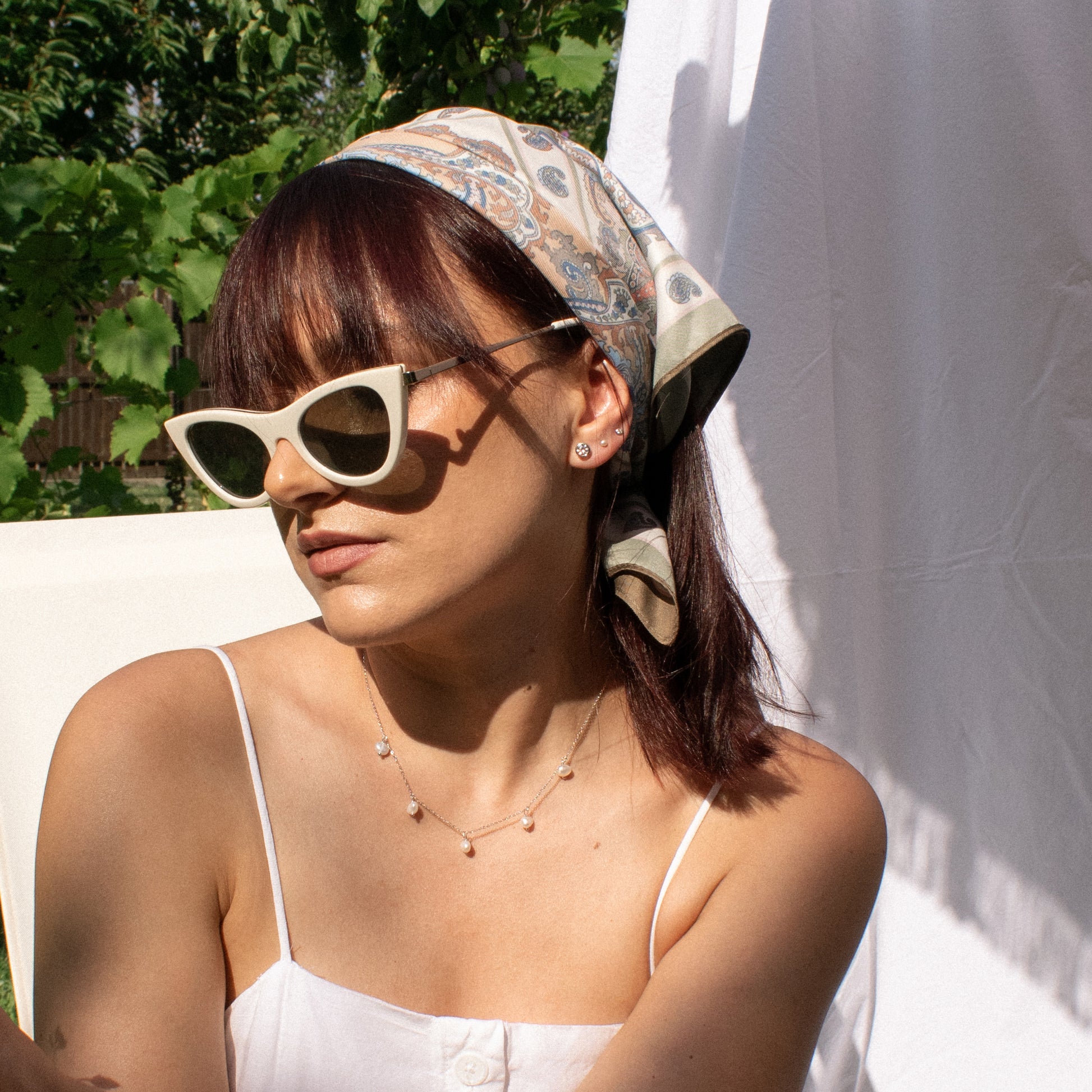 A woman wearing three silver-toned stud earrings, including one larger round clear-stone earring and two smaller studs, one with a small pearl and one with mini crystal . She is wearing a patterned scarf in warm natural sunlight.