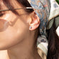 Close-up of a woman's ear wearing three silver-toned stud earrings, including one larger round clear-stone earring and two smaller studs, one with a small pearl and one with mini crystal . She is wearing a patterned scarf in warm natural sunlight.