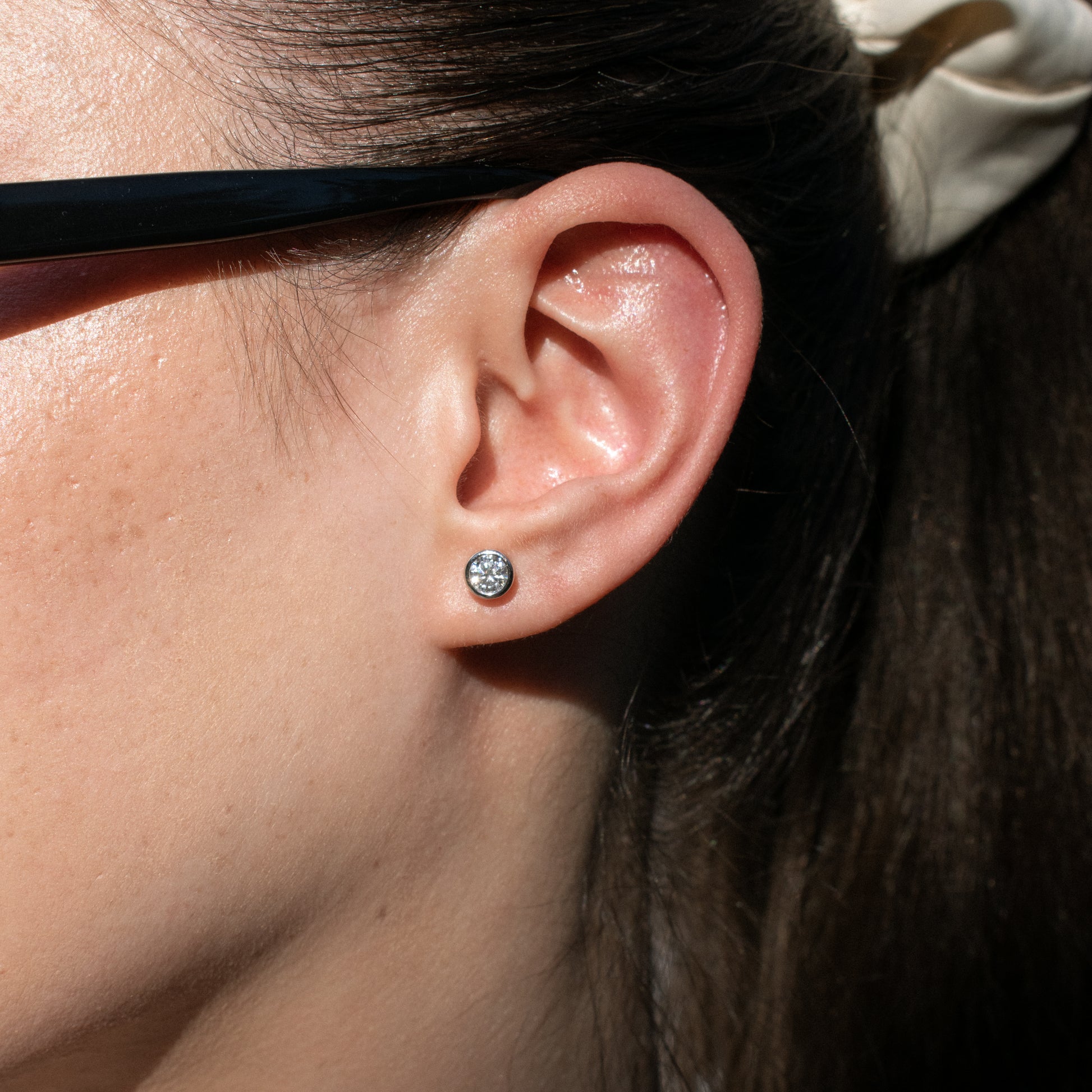 A close-up of a person's ear wearing a moissanite tennis round stud earring with a clear gemstone in a silver-toned setting. 