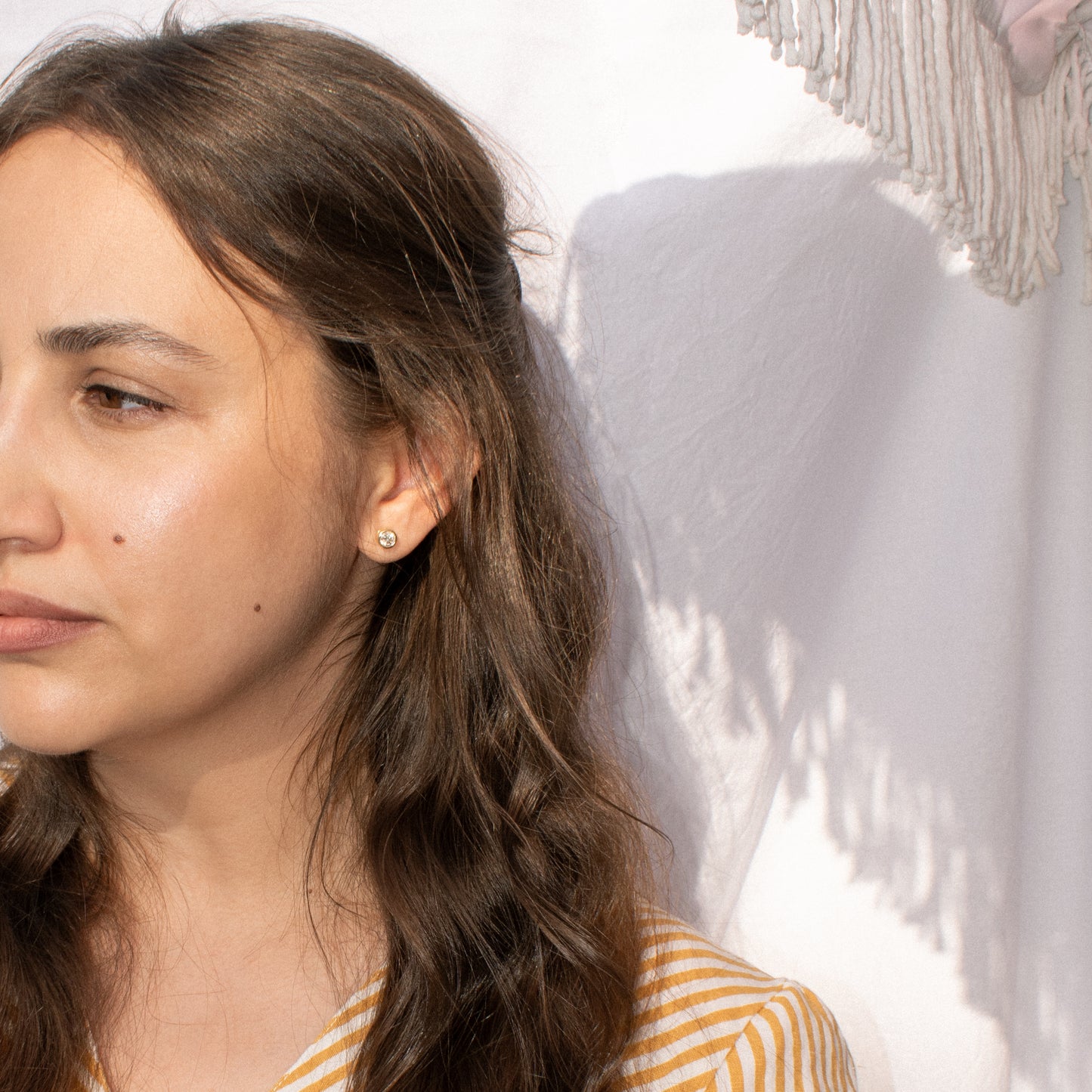 A close-up side profile of a woman with long brown hair wearing a moissanite gold stud earring, set against a white textured wall with soft shadows.