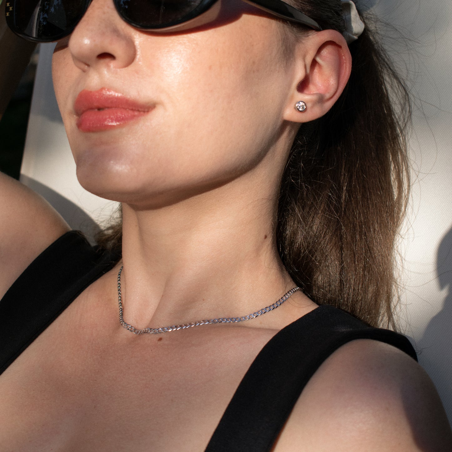 A close-up lifestyle shot of a woman wearing a moissanite silver stud earring, wearing a black top and sunglasses against a bright, sunlit background.