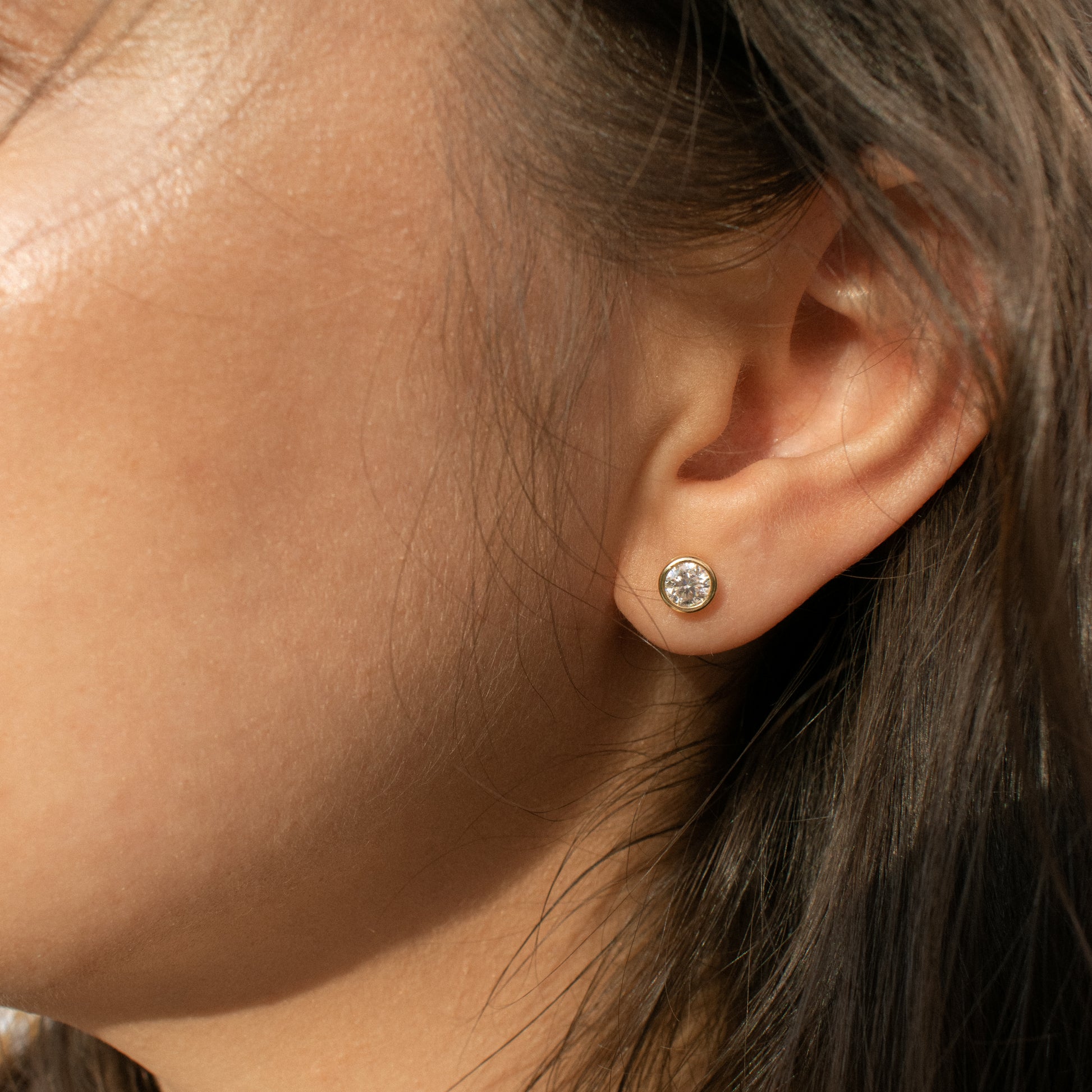 A close-up of a person's ear wearing a moissanite round stud earring with a textured silver-toned setting and a clear faceted stone.