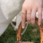 A close-up of a person's hand wearing two sterling silver rings, including one with a small  zirconia stone and one adjustable ring, holding a brown woven leather bag handle outdoors.