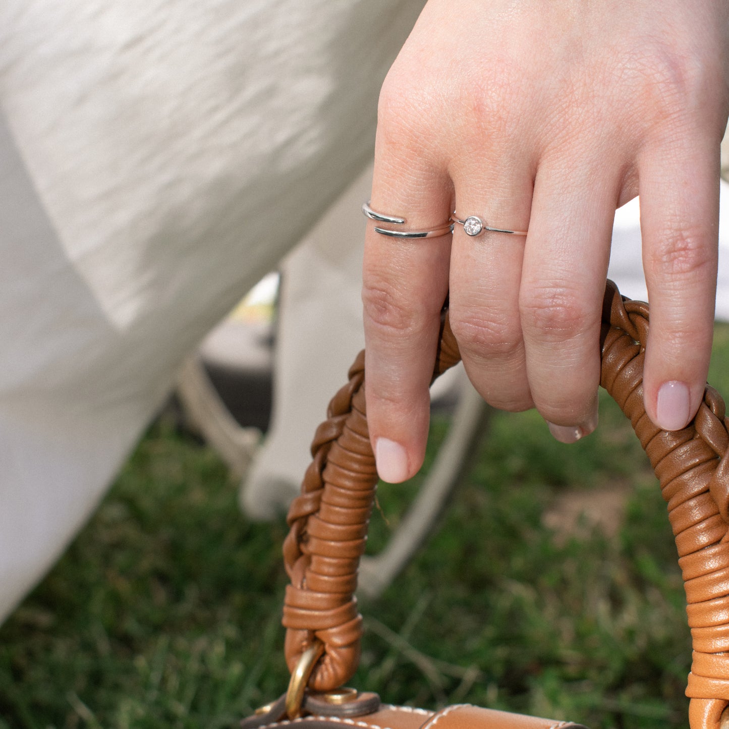 A close-up of a person's hand wearing two sterling silver rings, including one with a small  zirconia stone and one adjustable ring, holding a brown woven leather bag handle outdoors.