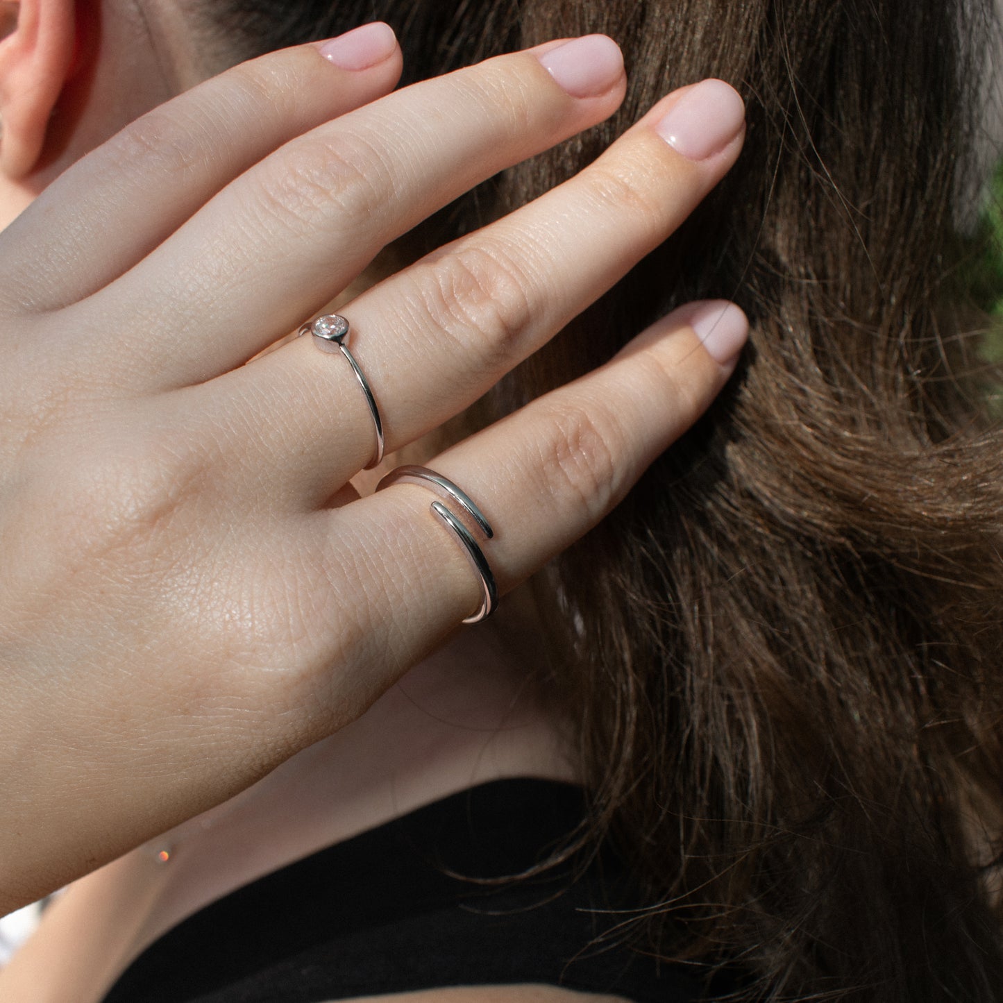 A close-up of a person's hand wearing two slim silver rings, one with a small round clear gemstone and one open-ended band, against dark brown hair.