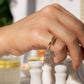 A close-up of a hand wearing a delicate adjustable double-band ring with two parallel gold-toned bands, positioned over white chess pieces in a softly blurred setting.