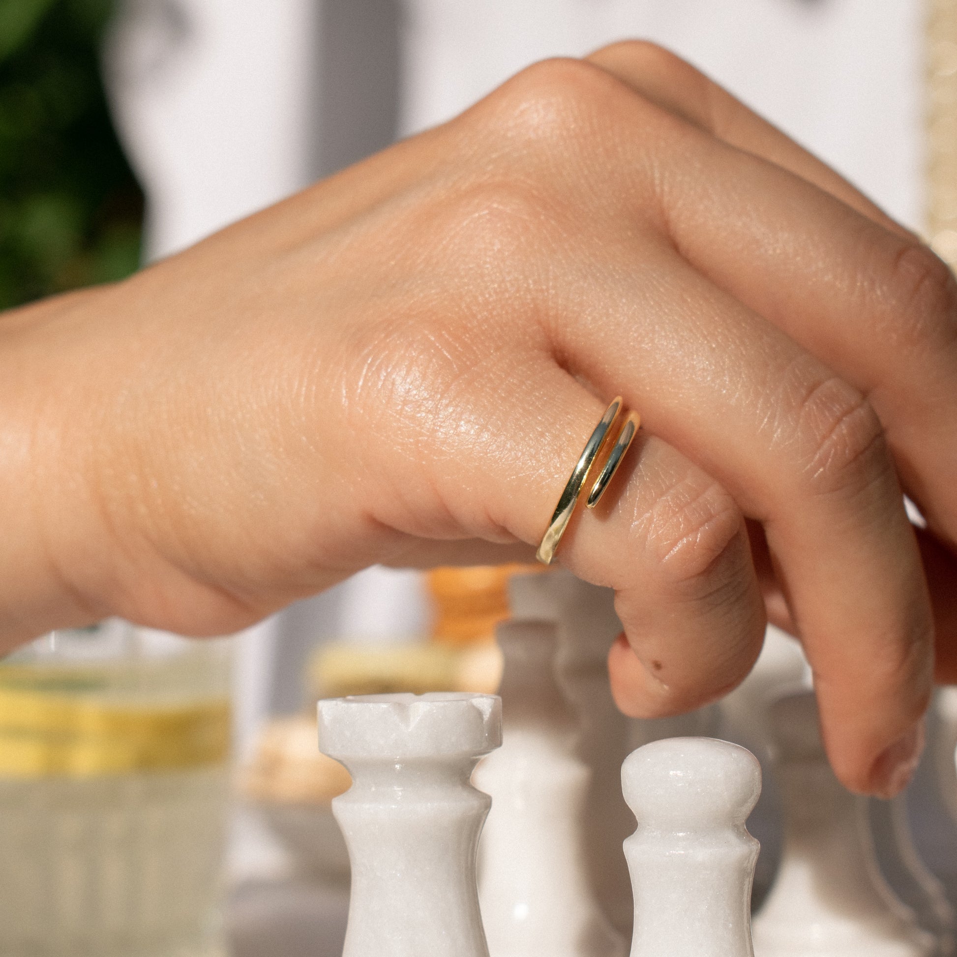 A close-up of a hand wearing a delicate adjustable double-band ring with two parallel gold-toned bands, positioned over white chess pieces in a softly blurred setting.