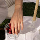 A close-up lifestyle shot of a person's hand resting on a glass bowl of berries, wearing a delicate gold chain bracelet on the wrist and a sterling silver gold plated ring with zirconia stone on the finger, set against an outdoor picnic scene with a white tablecloth, candle, and grass.
