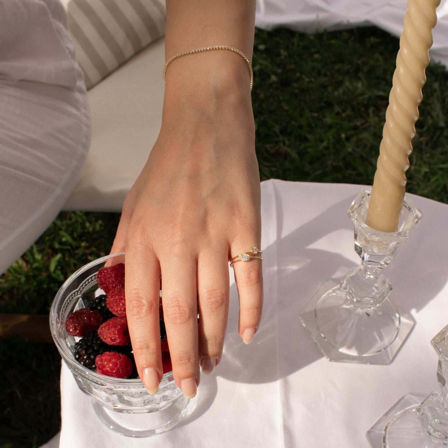 A close-up lifestyle shot of a person's hand resting on a glass bowl of berries, wearing a delicate gold chain bracelet on the wrist and a sterling silver gold plated ring with zirconia stone on the finger, set against an outdoor picnic scene with a white tablecloth, candle, and grass.