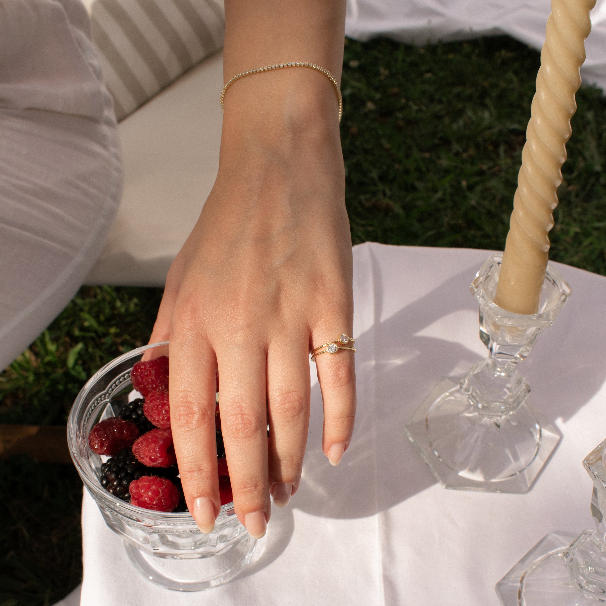 A close-up lifestyle shot of a person's hand resting on a glass bowl of berries, wearing a delicate gold chain bracelet on the wrist and a sterling silver gold plated ring with zirconia stone on the finger, set against an outdoor picnic scene with a white tablecloth, candle, and grass.