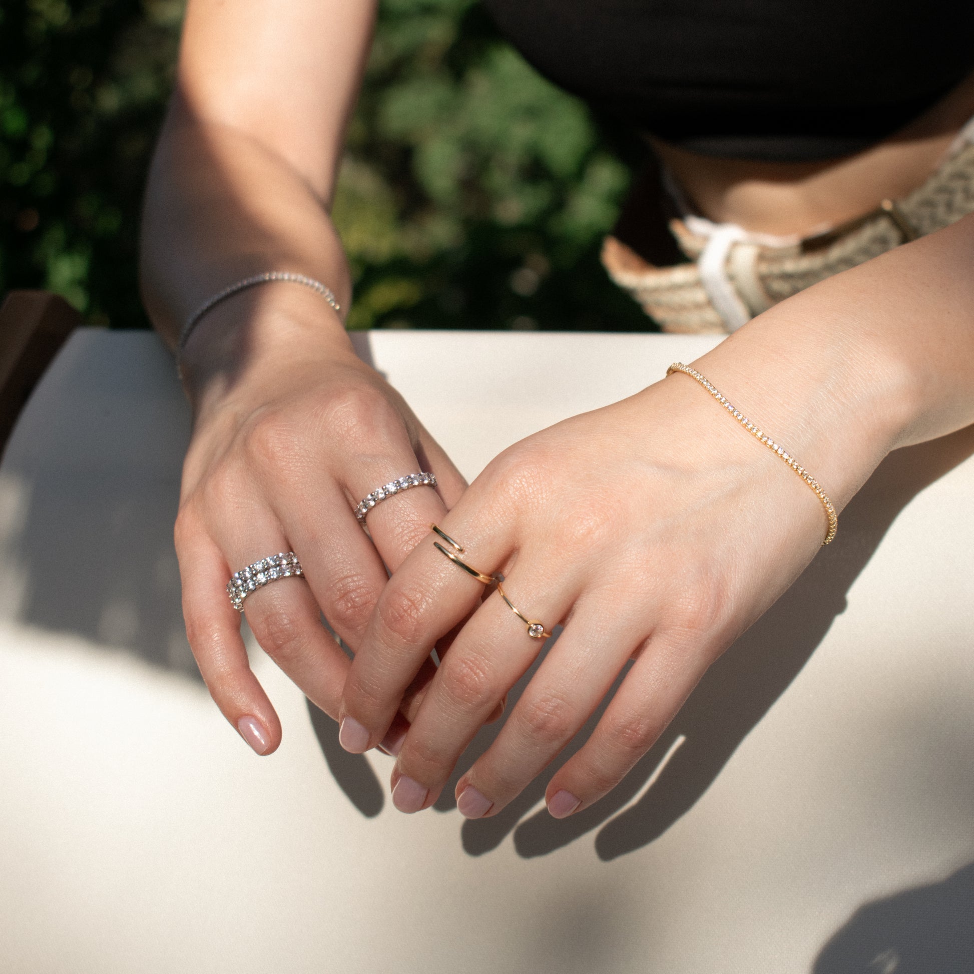 A close-up of a person's hands wearing delicate jewelry, including silver tennis rings with zirconia stones, a gold ring, a gold tennis bracelet, and a silver tennis bracelet, set against a bright outdoor background with soft green foliage.