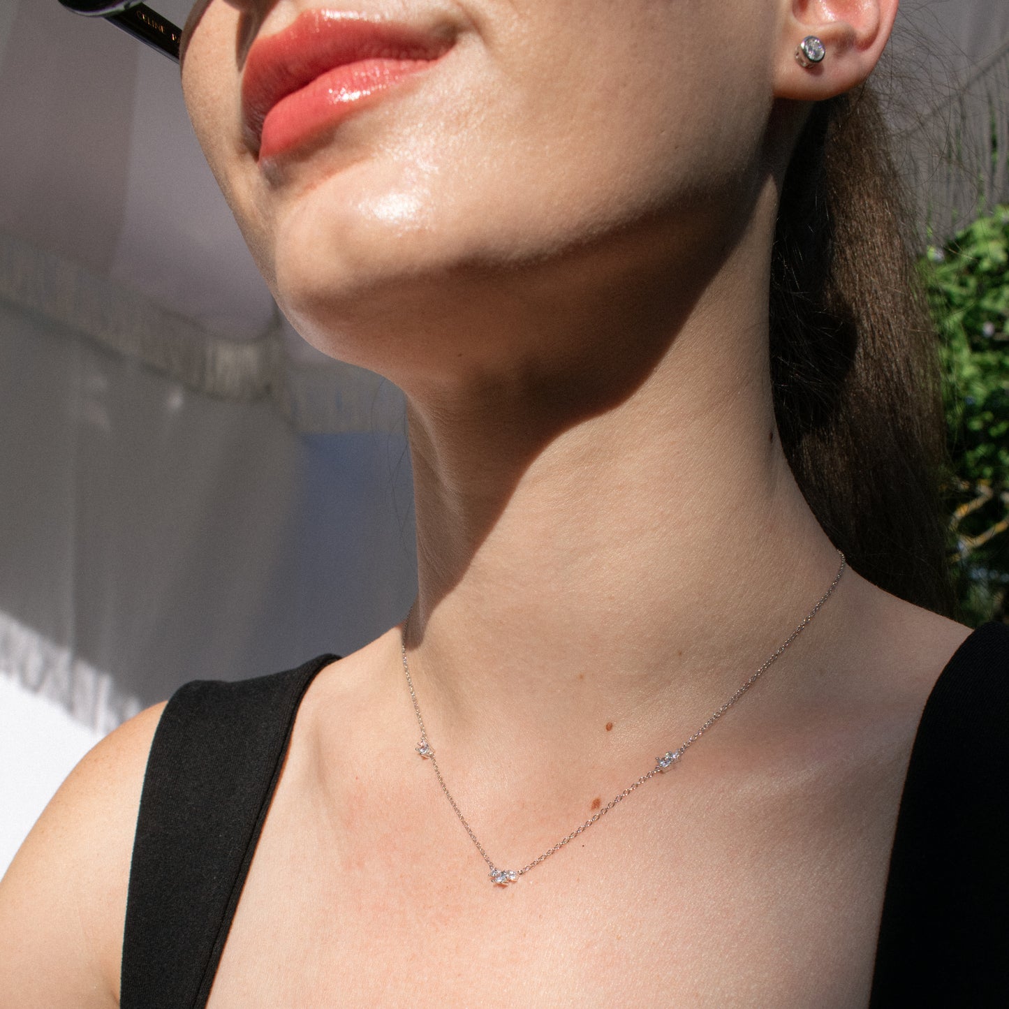 A close-up lifestyle shot of a woman wearing a delicate silver necklace with small star-shaped clear gemstones and a matching stud earring, shown in bright natural sunlight.