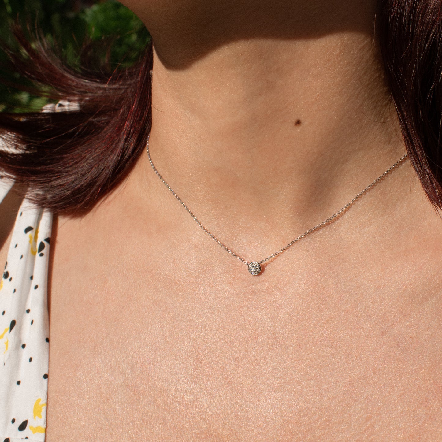 A close-up of a delicate silver chain necklace with a small, round, clear zirconia stones pendant worn by a person outdoors.