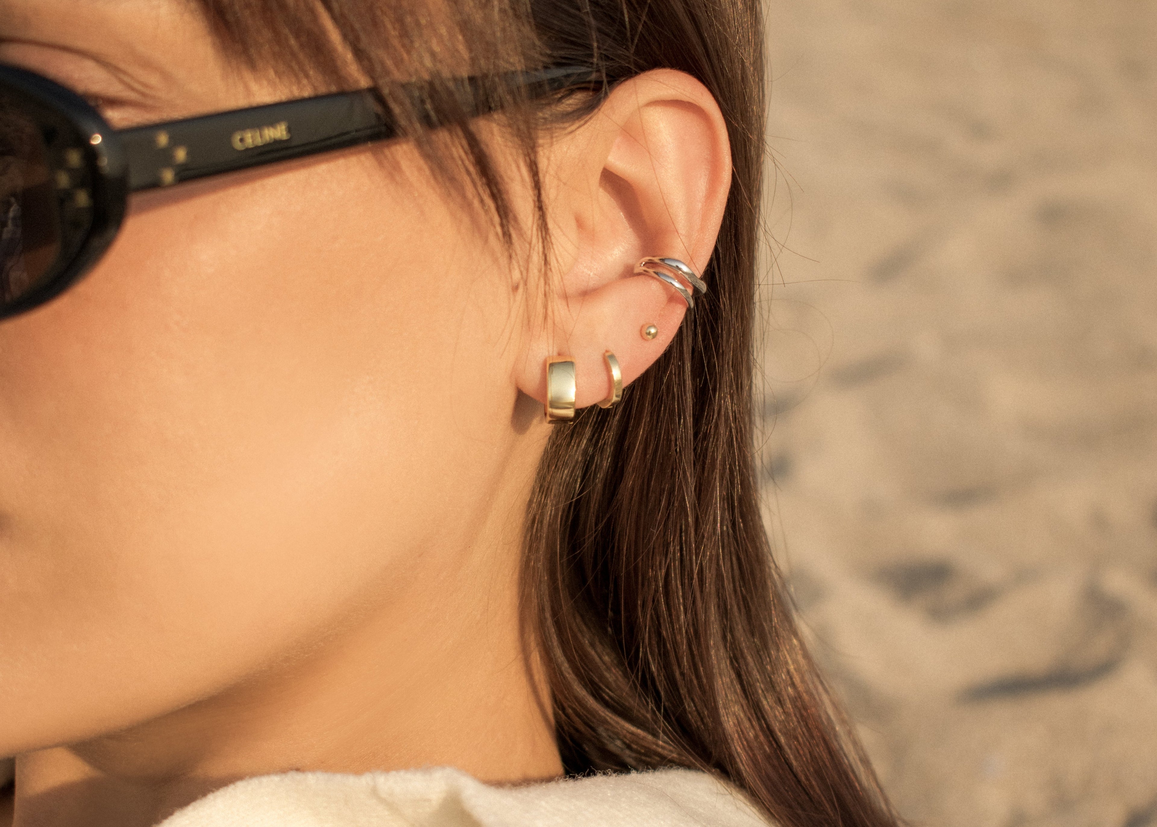 Cadru apropiat pe urechea unei femei care poarta cercei din argint unii placati cu rodiu si unii placati cu aur 18k vermeil pe fundal blurat. Close-up of a person ear wearing silver earrings and cuff earrings with a blurred background