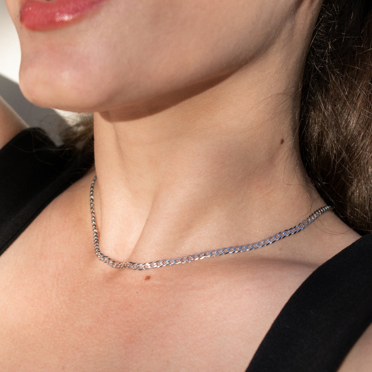A close-up of a woman's neck and upper chest wearing a silver diamond cut cuban chain necklace with a textured, woven-like pattern.