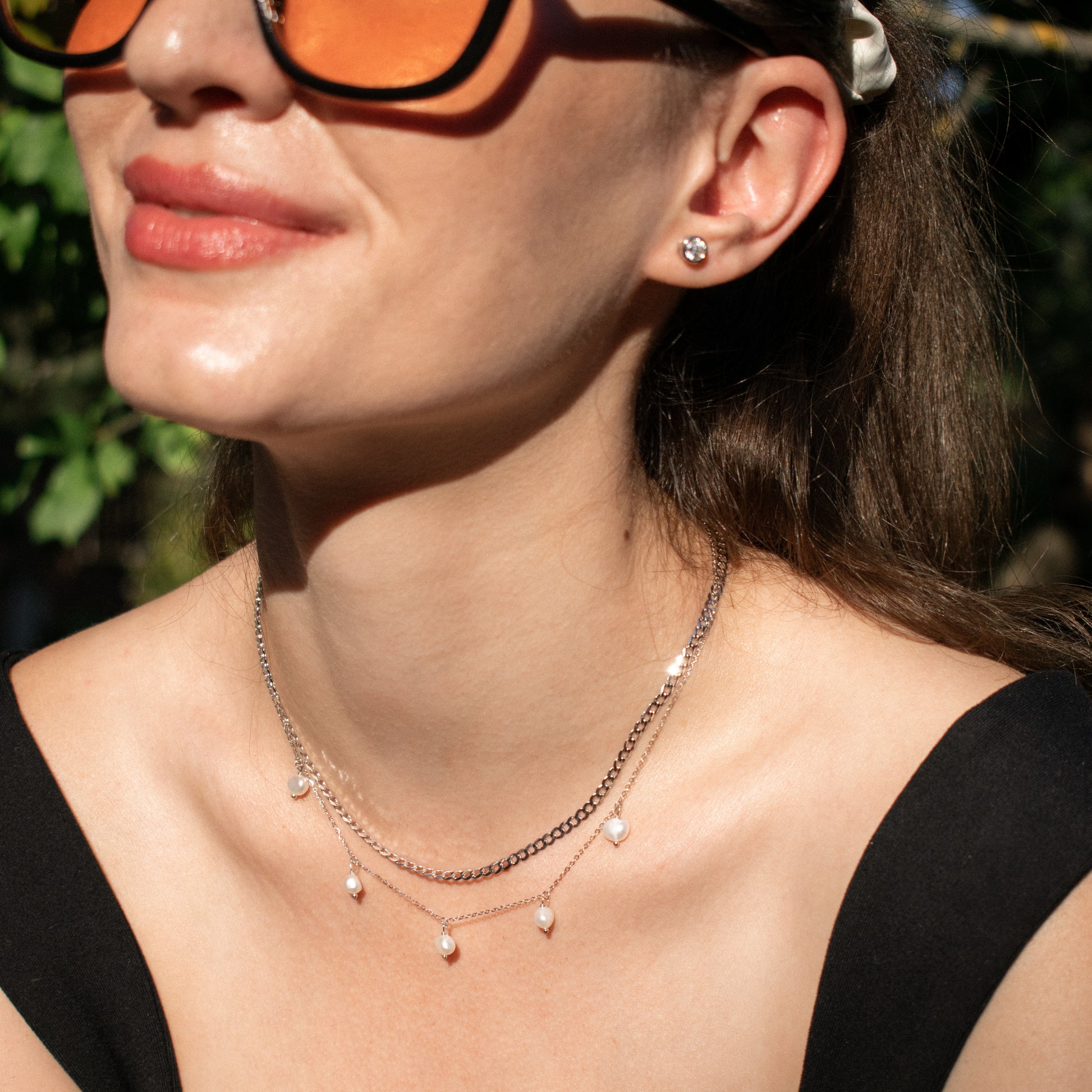 A close-up lifestyle shot of a woman wearing a layered necklace with small white pearl-like drops on a silver diamond cut cuban chain, along with a stud earring and orange-tinted sunglasses.